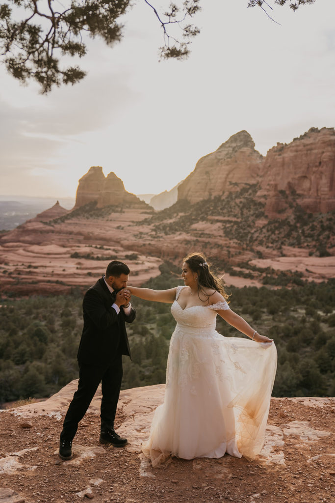 groom kissing bride's hand with red rock mountains in the background at merry go round rock in sedona, arizona