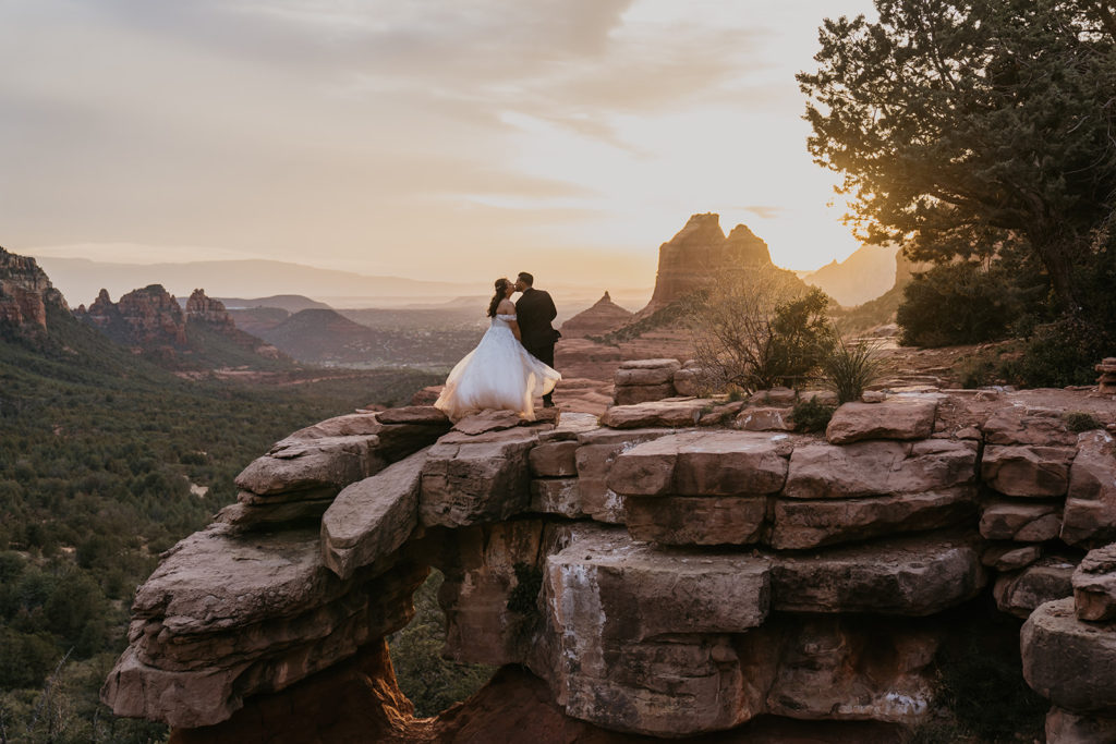 Bride and groom kissing with the sunset in the background on red rocks at merry go round rock in sedona, arizona
