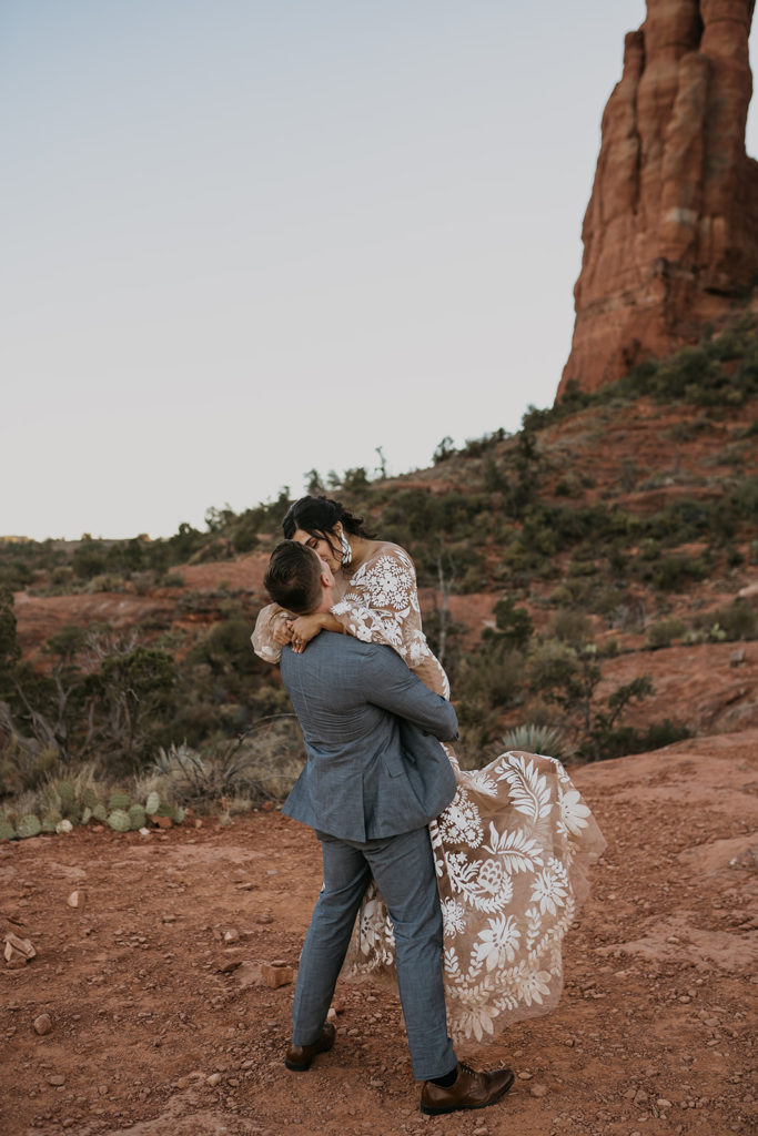 groom picking up bride and kissing with a red mountain in the background at their elopement at cathedral rock in sedona