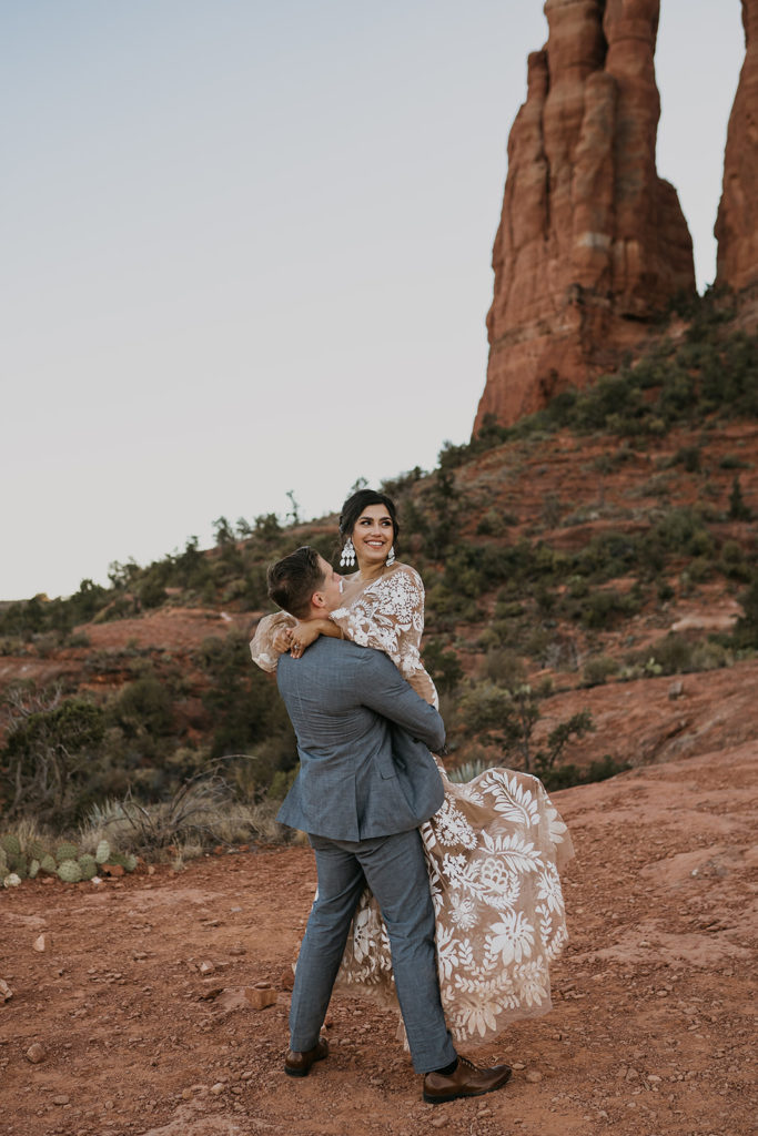 groom picking up bride and kissing with a red mountain in the background at their elopement at cathedral rock in sedona