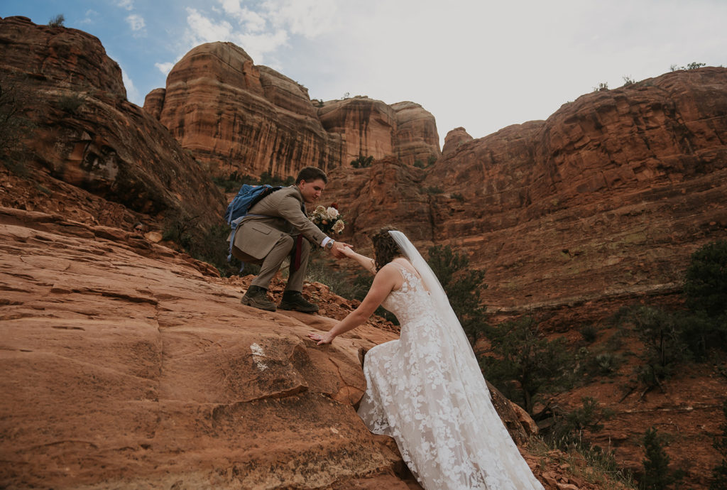groom helping bride in wedding dress up on a mountain